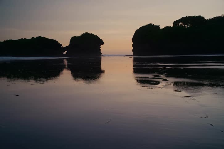 A tranquil beach at twilight features silhouetted rock formations and their perfect reflection on the wet sand
