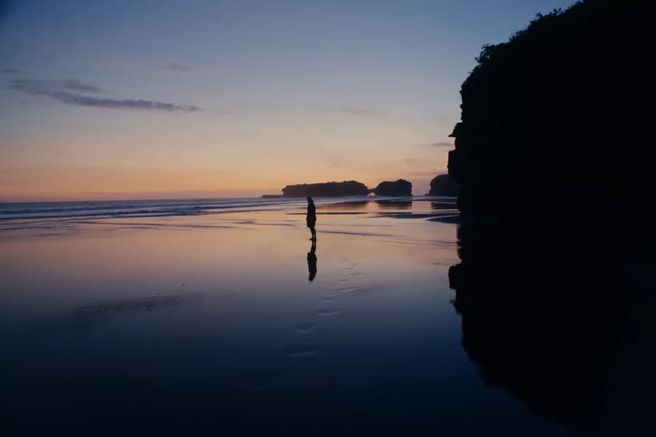 A serene wide shot of a silhouetted person standing alone on a calm, reflective beach during a beautiful twilight glow