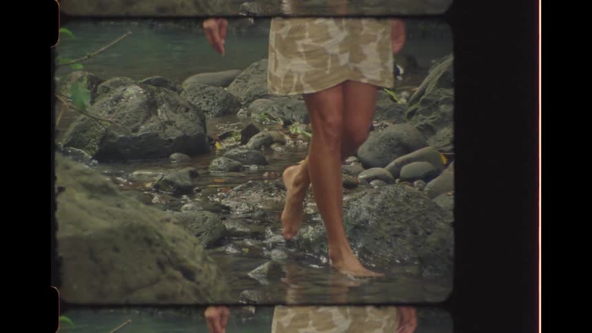 A woman walks barefoot in slow motion through a rocky jungle stream in vintage-style footage