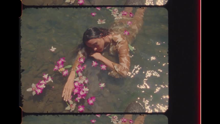 Vintage film-style footage of a serene woman in a dress floating in sunlit water amongst pink and white orchid flowers