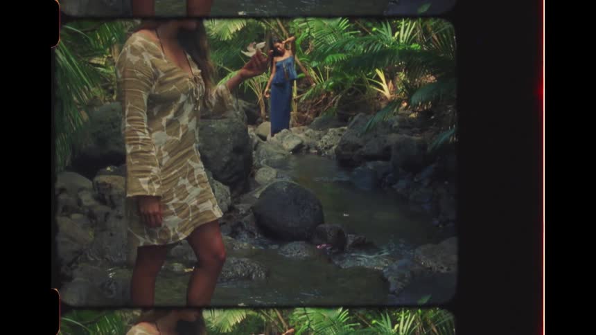With a vintage film aesthetic, a woman gathers flowers from a rocky stream in a lush tropical jungle while another woman stands in the background