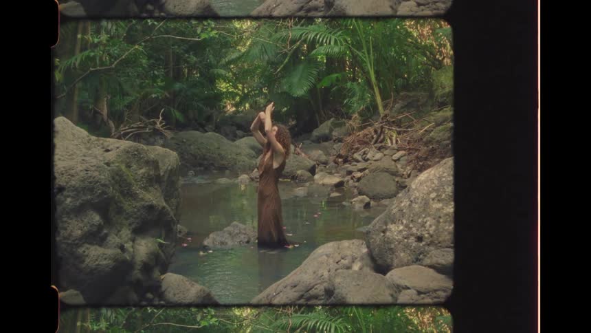 Vintage-style footage of a woman in a long flowing dress gracefully dancing in a secluded jungle stream surrounded by rocks and lush greenery