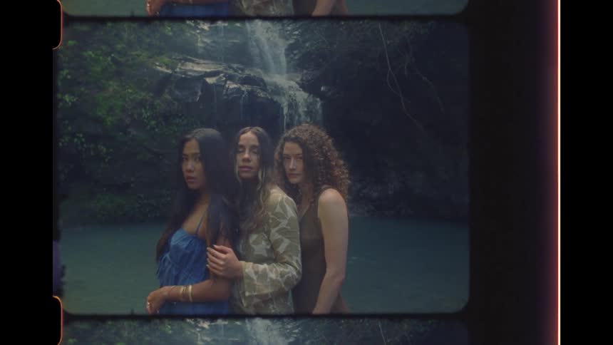 Three diverse and ethereal women pose together in a jungle lagoon in front of a cascading waterfall, captured on vintage film