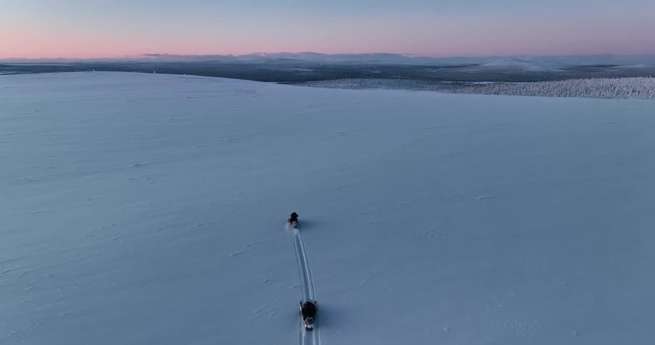 An aerial view of two snowmobiles driving away from the camera across a vast and empty snowy landscape during a pastel-colored sunset