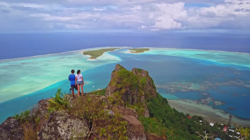 A drone ascends to reveal a couple standing on a mountain summit, admiring the spectacular view of a tropical island paradise