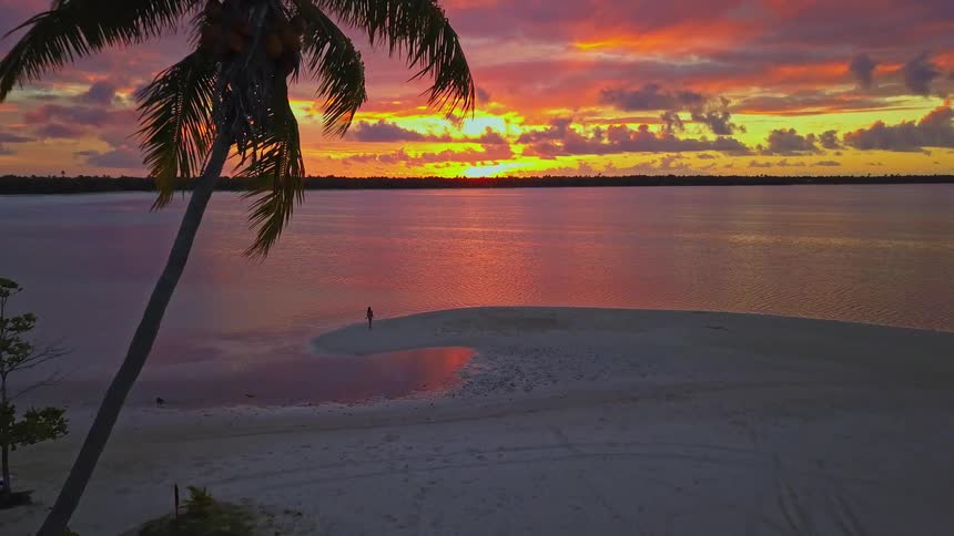 An aerial view captures a lone figure on a tropical sandbar, silhouetted against a spectacular fiery sunset reflected in the calm ocean