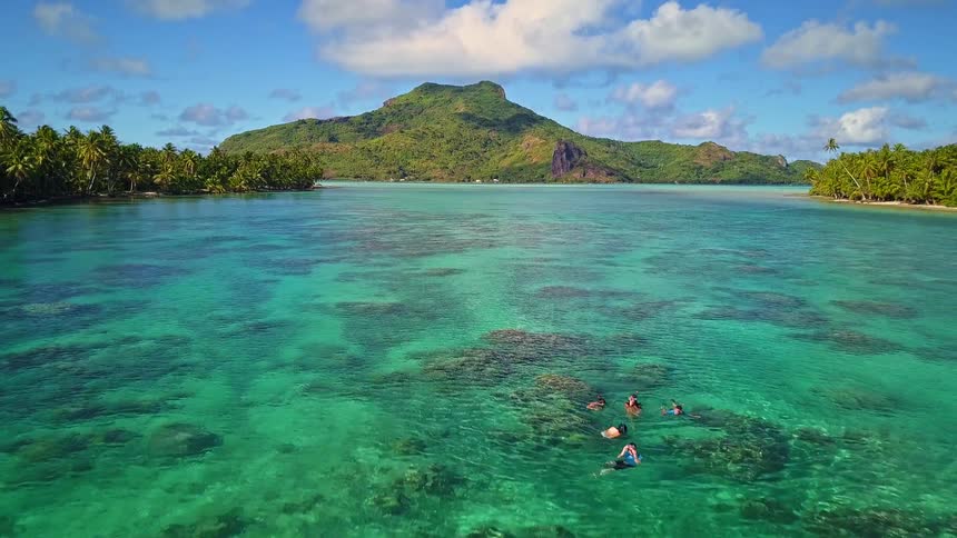 A stunning aerial view shows a group of people snorkeling in the vibrant turquoise waters of a tropical island lagoon, with a majestic green mountain in the distance