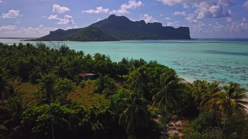 An aerial shot slowly ascends over a vibrant green tropical island, showcasing the turquoise water and a beautiful mountain in the background