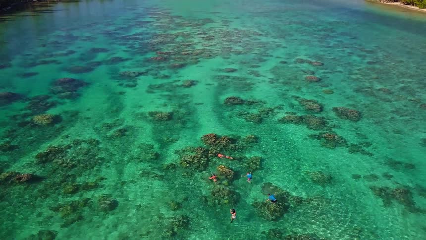 An aerial view pulls back from people snorkeling in a clear turquoise lagoon to reveal the stunning tropical islands and mountains of Bora Bora