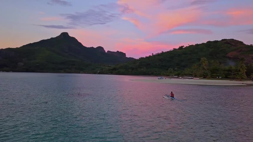 A lone person paddles an outrigger canoe across the tranquil turquoise water of a tropical island bay during a vibrant sunset