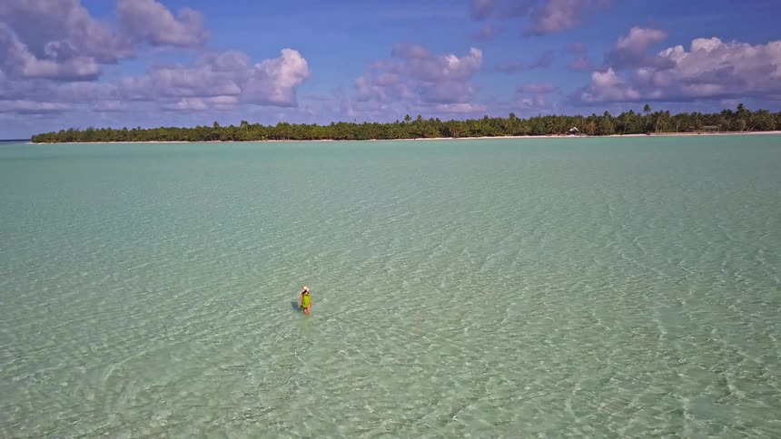 An adventurous traveler with a backpack wades through a vast, serene turquoise lagoon towards a remote tropical island