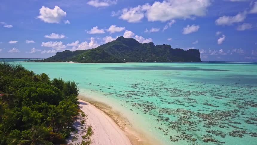 Ascending aerial shot reveals a pristine white sand beach, a turquoise lagoon, and a lush mountainous island under a beautiful blue sky