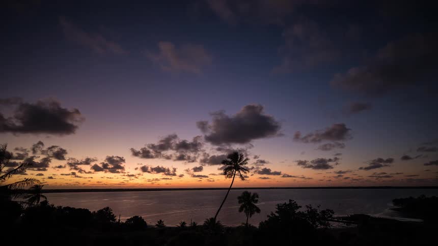 A beautiful timelapse of clouds moving over a tropical bay during a colorful sunset with silhouetted palm trees