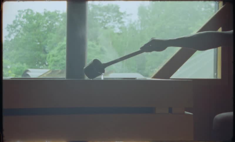 A person's arm pours water with a ladle onto hot stones to create steam in a sauna as it rains outside the window