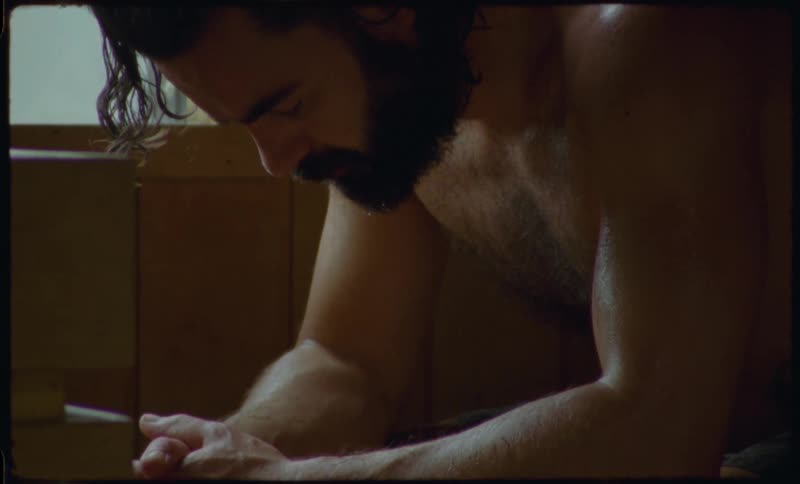 A bearded man meditates in a hot wooden sauna as sweat drips from his face in a grainy, vintage-style clip