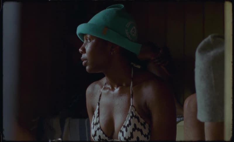 A woman in a bikini and sauna hat drinks water to rehydrate while sitting in a hot, steamy wooden sauna