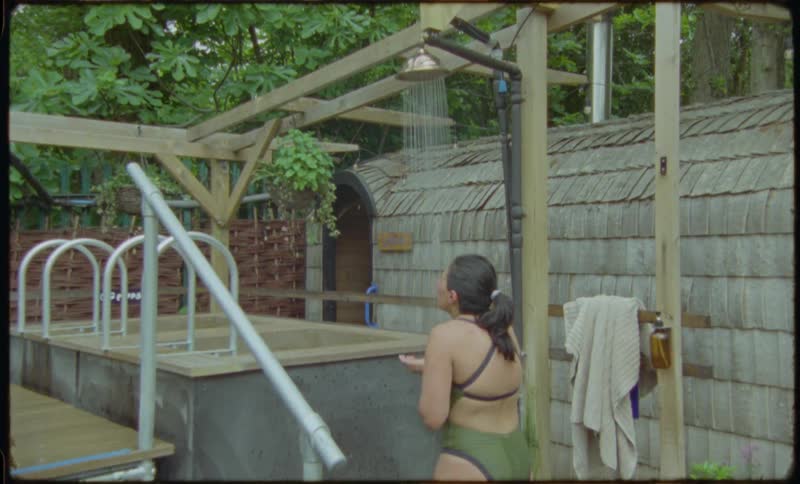 A woman in a swimsuit enjoys a refreshing outdoor shower at a rustic spa retreat surrounded by lush greenery
