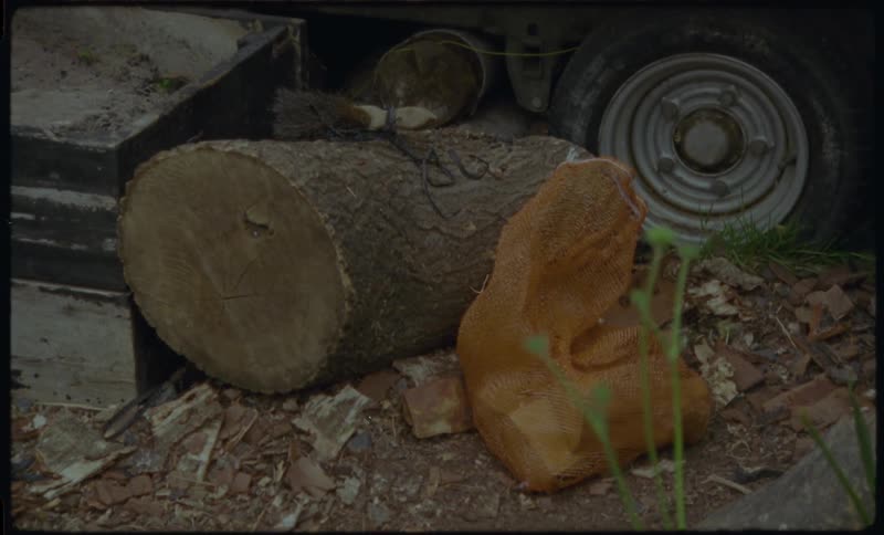 A vintage, static shot shows a chopped log and a mesh bag of potatoes resting amongst rubble and junk next to a vehicle's wheel