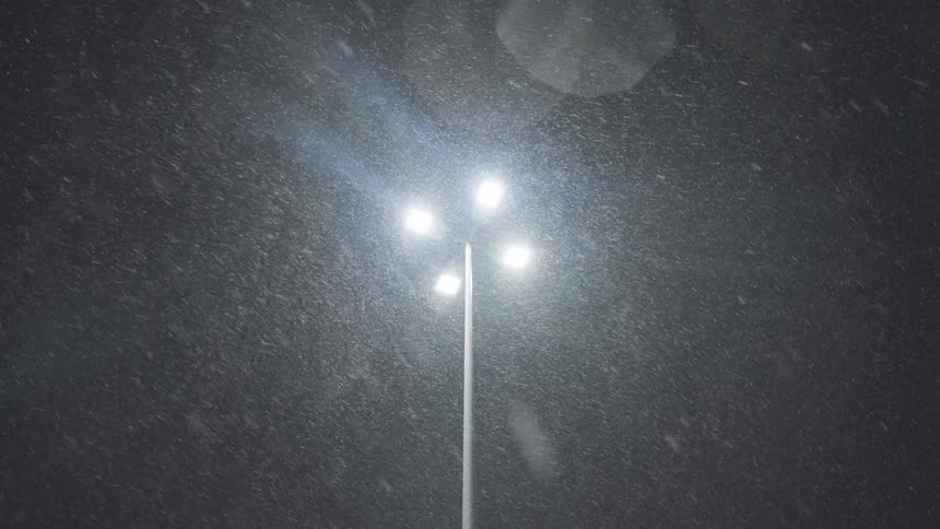 Heavy snow falls at night, illuminated by the bright glow of a street lamp