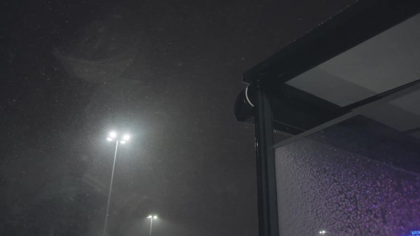 Heavy snow falls at night, illuminated by bright streetlights next to a bus shelter