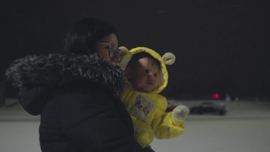 A mother holds her baby in a yellow bear snowsuit as they experience their first snowfall together on a cold winter night