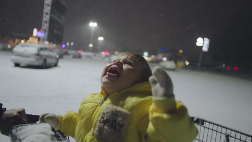 A happy baby in a yellow snowsuit laughs hysterically in slow motion while riding in a shopping cart as snow falls at night