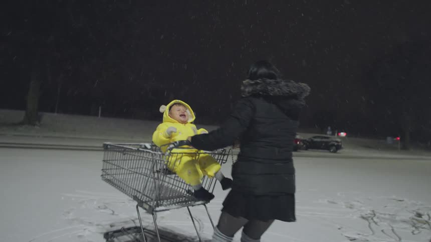 A laughing mother pushes her toddler in a shopping cart through a snowy parking lot at night in super slow motion
