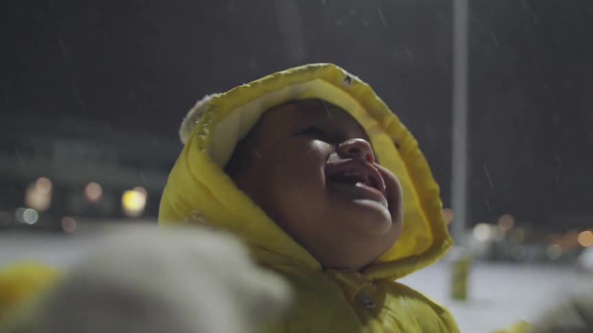 An adorable baby in a yellow snowsuit looks up at the sky with pure joy, trying to catch snowflakes on its tongue during a magical winter night