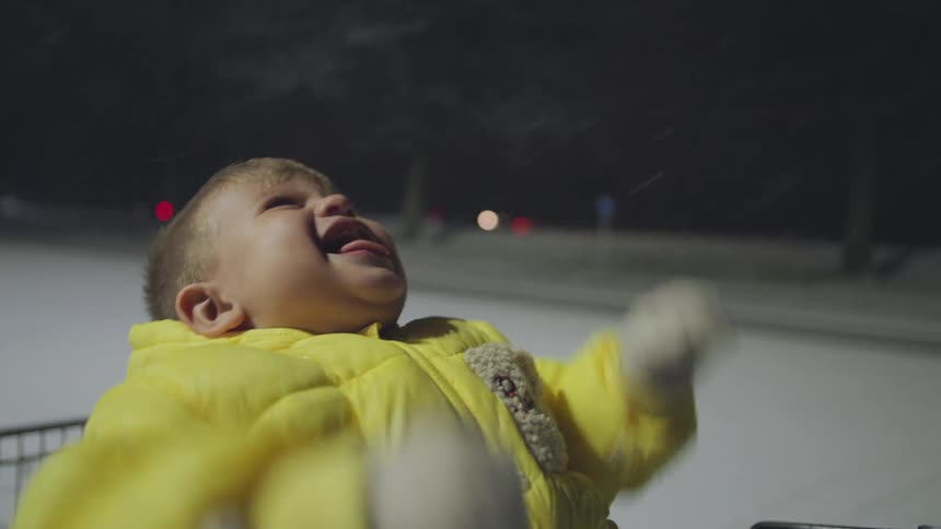 A bundled-up toddler in a yellow coat transitions from crying to laughing with joy while catching snowflakes on their tongue on a winter night