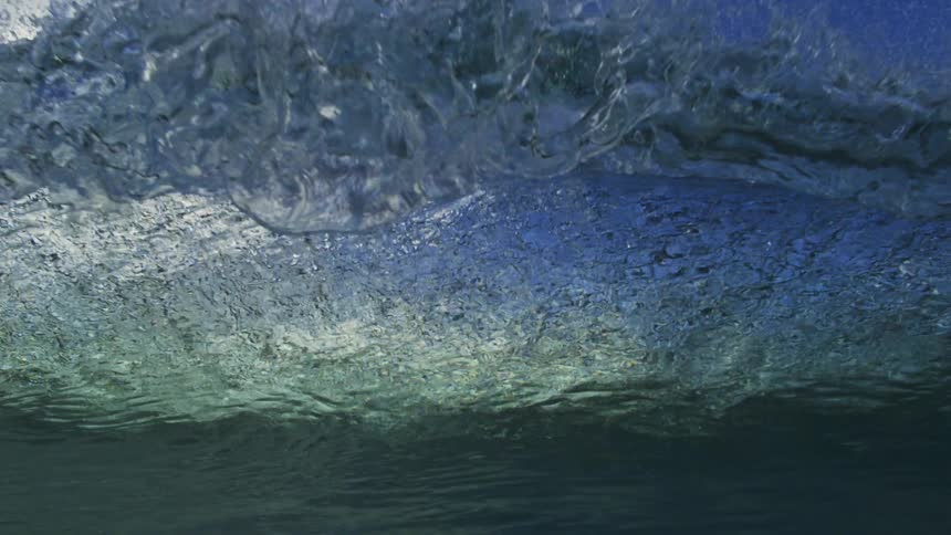An ocean wave crests against a cloudy blue sky before the camera is submerged underwater in super slow motion