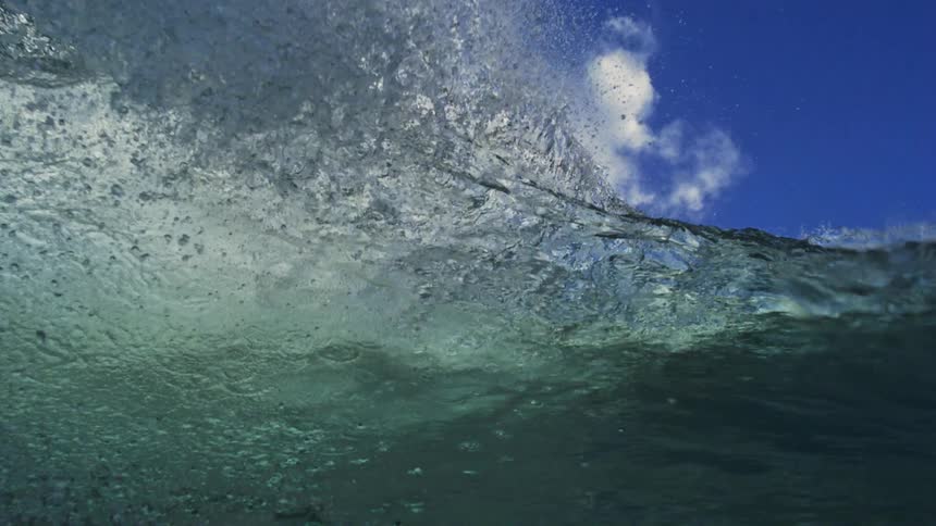 Slow motion over-under shot shows a powerful ocean wave cresting against a blue sky before the camera submerges into the turbulent water