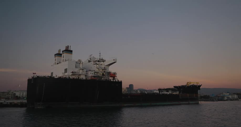 A large industrial cargo ship is docked in a calm harbor with a city skyline visible in the background during a colorful sunset