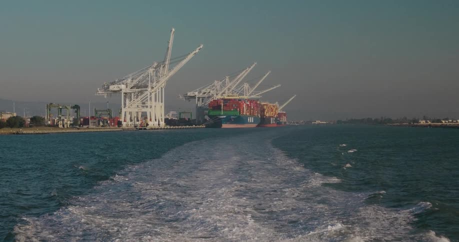 View from the back of a boat leaving an industrial shipping port with large cranes and cargo ships, with the boat's wake visible on the water
