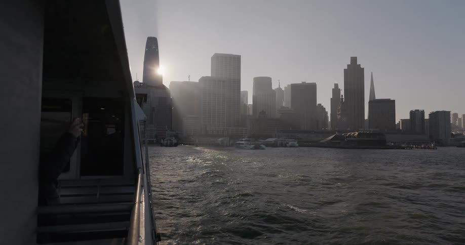A person takes a photo of the hazy San Francisco skyline from the side of a moving ferry as the sun shines brightly over the city