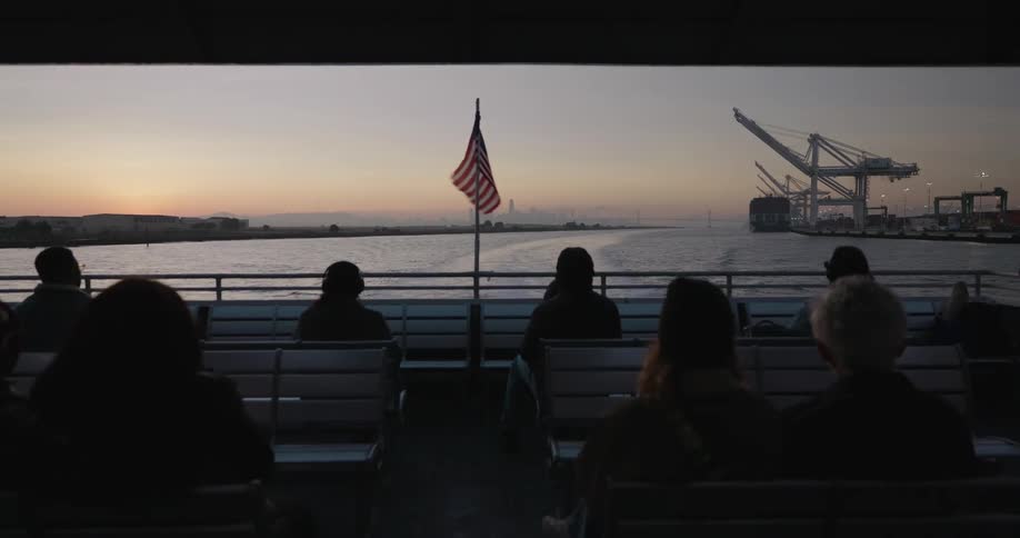 Silhouetted passengers on a ferry enjoy the view of an industrial port and distant city skyline at sunset