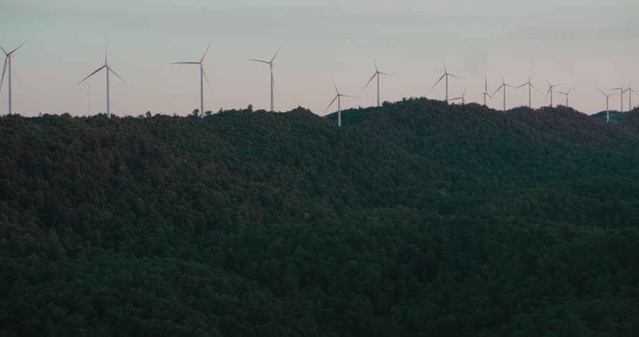 A long line of wind turbines spin peacefully along a forested mountain ridge at dusk