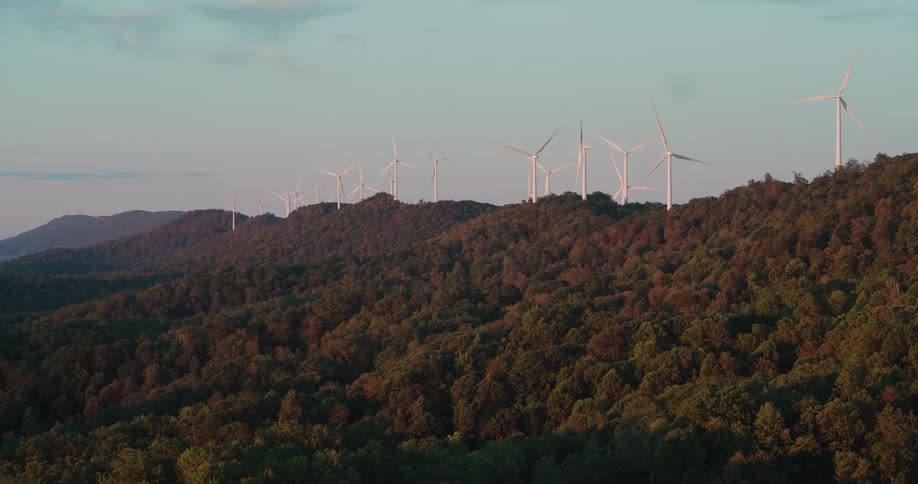 Wind turbines spin along a forested mountain ridge, harnessing clean energy as the warm light of sunset illuminates the landscape