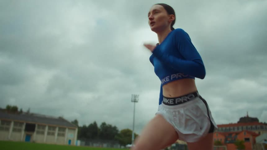 Slow-motion side view of a determined female athlete in athletic gear running on a track under an overcast sky