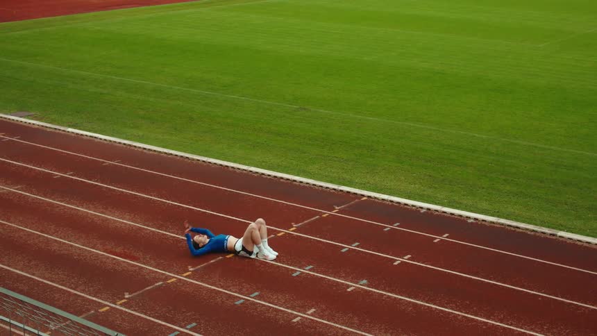 A tired female athlete gracefully falls onto a stadium running track to rest in slow motion