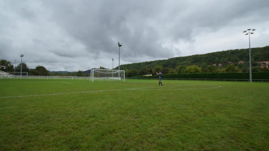 A soccer player practices corner kicks alone on an empty green field under a cloudy, overcast sky