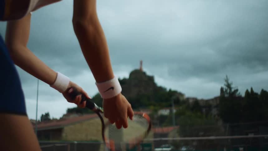 A female tennis player serves the ball in slow motion, captured from a dramatic low angle against a cloudy sky