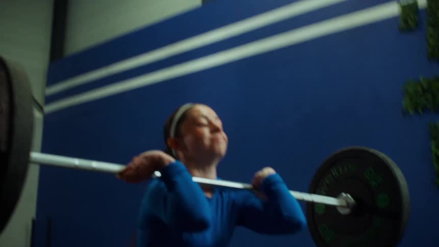 A strong, focused young woman performs a powerful barbell clean lift in slow motion at the gym