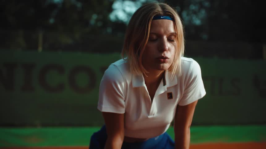 A determined female tennis player catches her breath, leaning on her racket in exhaustion on a clay court