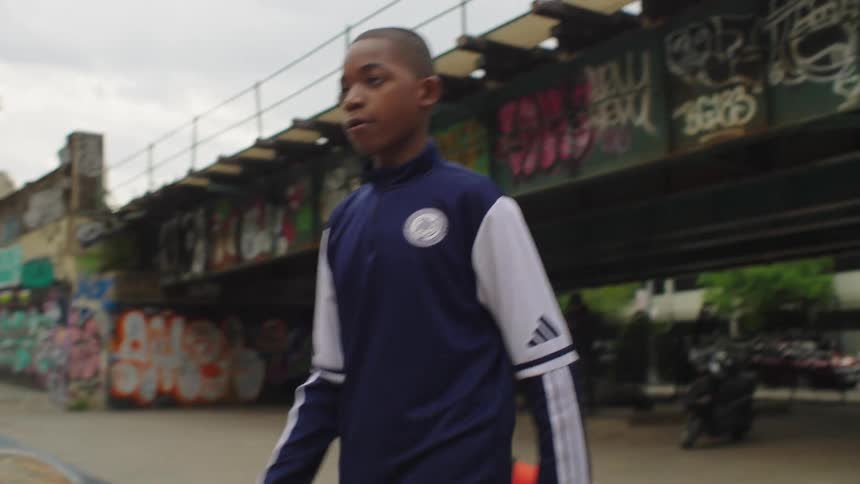 In slow motion, a focused young boy juggles a soccer ball in a graffiti-covered urban park