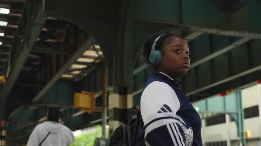 A thoughtful young Black woman holds a soccer ball and listens to music on her headphones while waiting on a city street corner