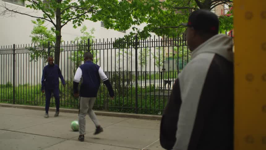A man watches two young men play soccer on a city sidewalk in slow motion with a rack focus