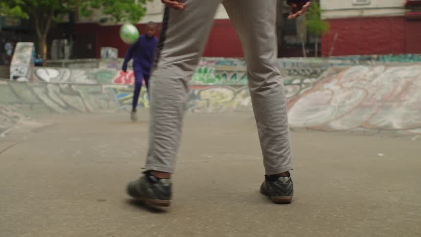 Two men practice their soccer skills, juggling and passing a ball in a gritty urban skatepark covered in graffiti
