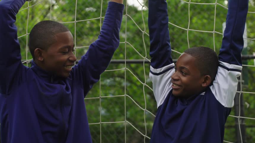A close-up shot of two young African American twin brothers smiling and talking as they playfully hang from a soccer goal