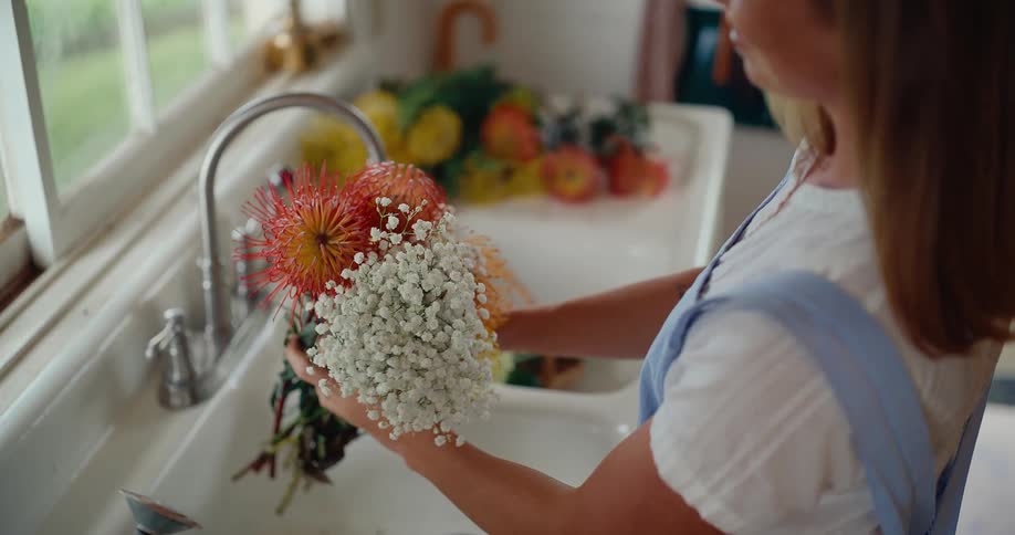 A woman in an apron gently rinses a vibrant bouquet of pincushion protea and baby's breath in a rustic farmhouse kitchen sink