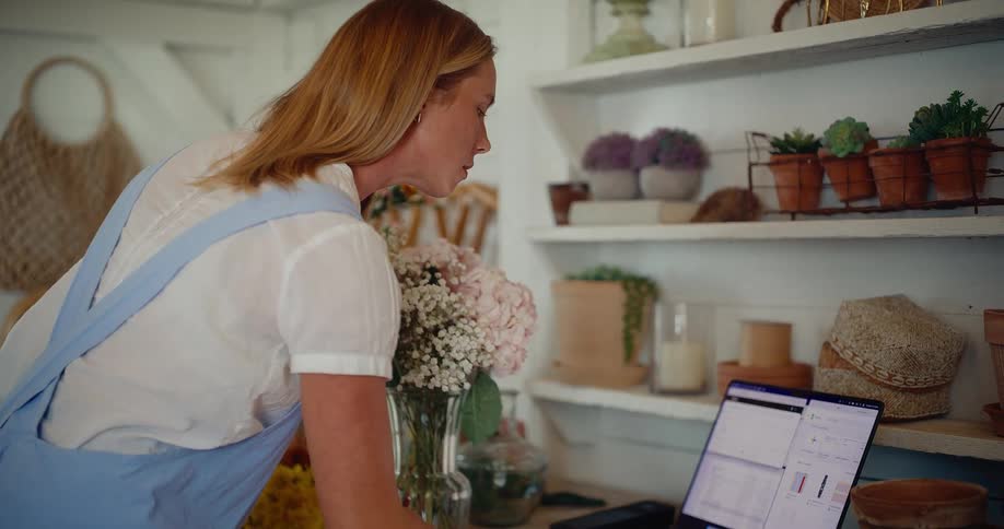 A female florist arranges a beautiful bouquet of flowers in her charming workshop before managing her small business on a laptop